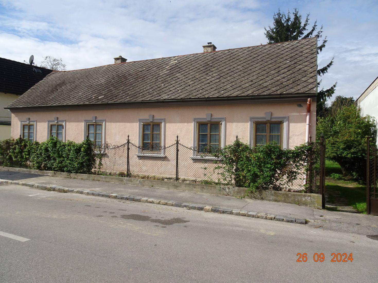 Einfaches Einfamilienhaus mit rosa Fassade, Ziegeldach und einem kleinen Vorgarten zur Straße hin.