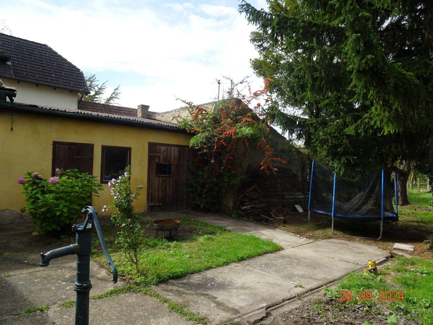Altes Nebengebäude mit Holztüren, einem Trampolin und einer Wasserpumpe im Gartenbereich.