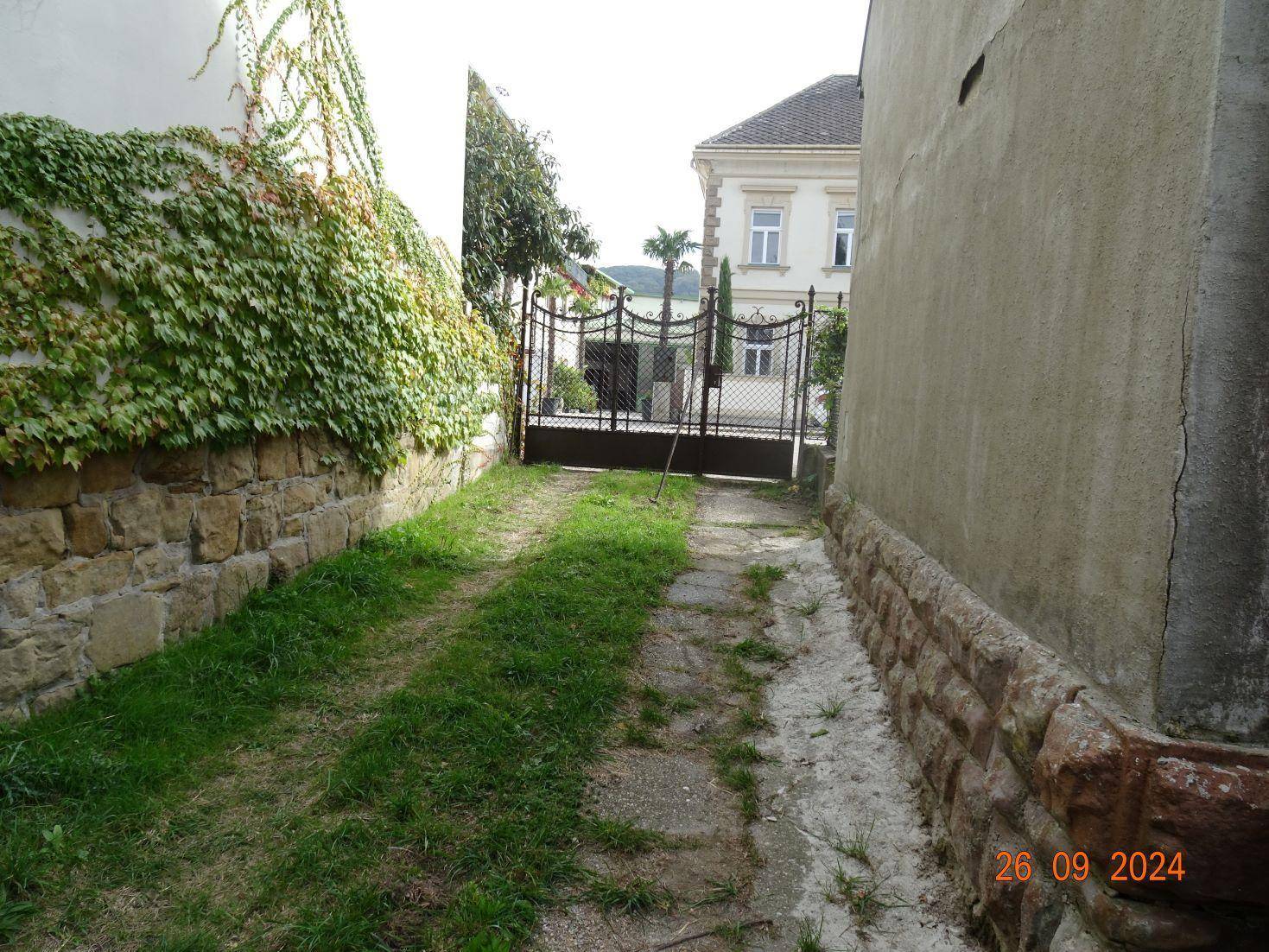 Schmaler Zugangsweg zwischen Gebäuden, gesäumt von Steinmauern und Efeu, führt zu einem Tor.