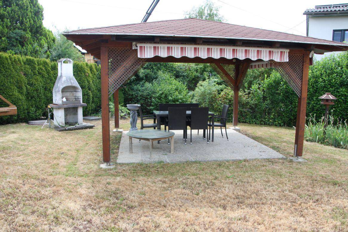 Überdachter Sitzbereich im Garten mit Grillstelle und grüner Hecke im Hintergrund.