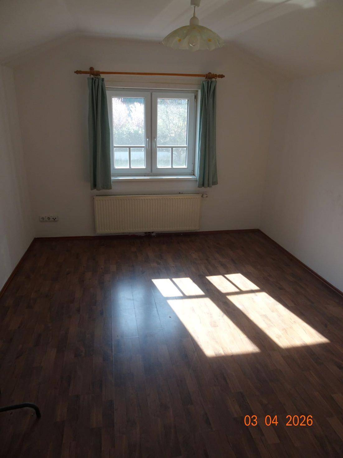 Leerer Raum mit Laminatboden und einem Fenster, das Sonnenlicht auf den Boden wirft.