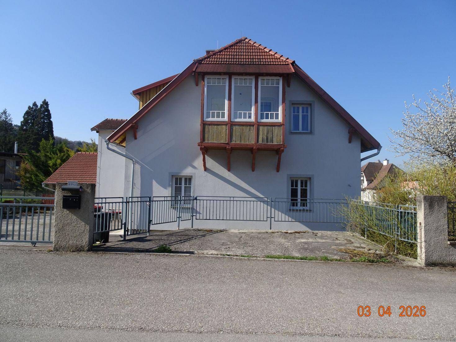 Vorderansicht des Hauses von der Straße mit einem kleinen Erker und Metallzaun.