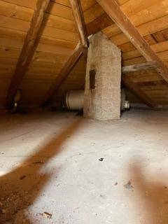 Dachboden mit sichtbaren Holzbalken, schräger Holzdecke und einem zentralen gemauerten Schornstein.