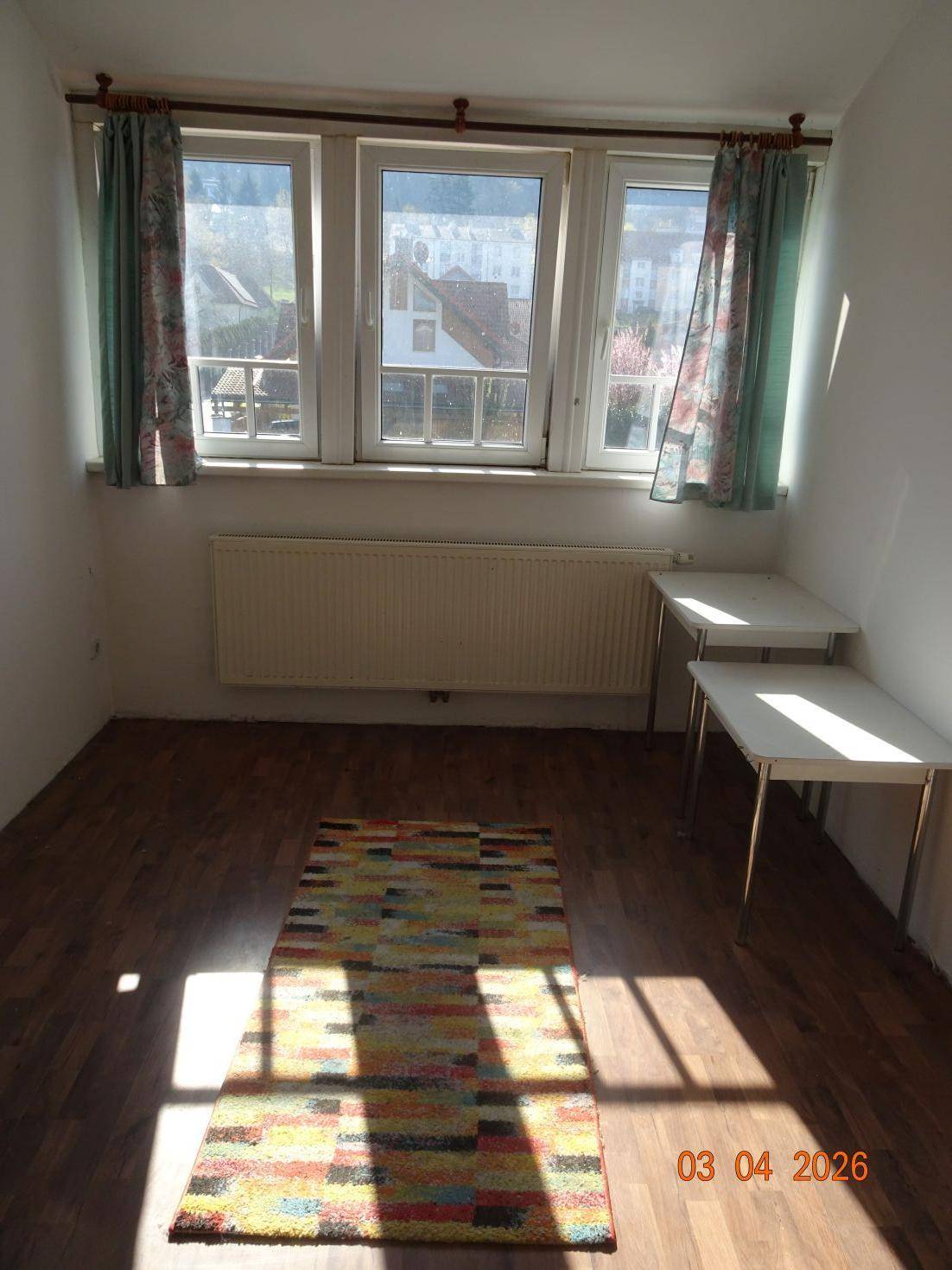 Lichtdurchfluteter Raum mit mehreren Fenstern und einem bunten Teppich auf dem Laminatboden.