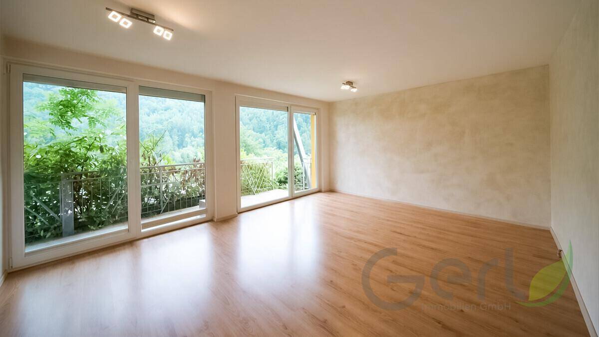 Leeres Wohnzimmer mit hellem Holzboden und großen Fenstern mit Zugang zum Balkon.