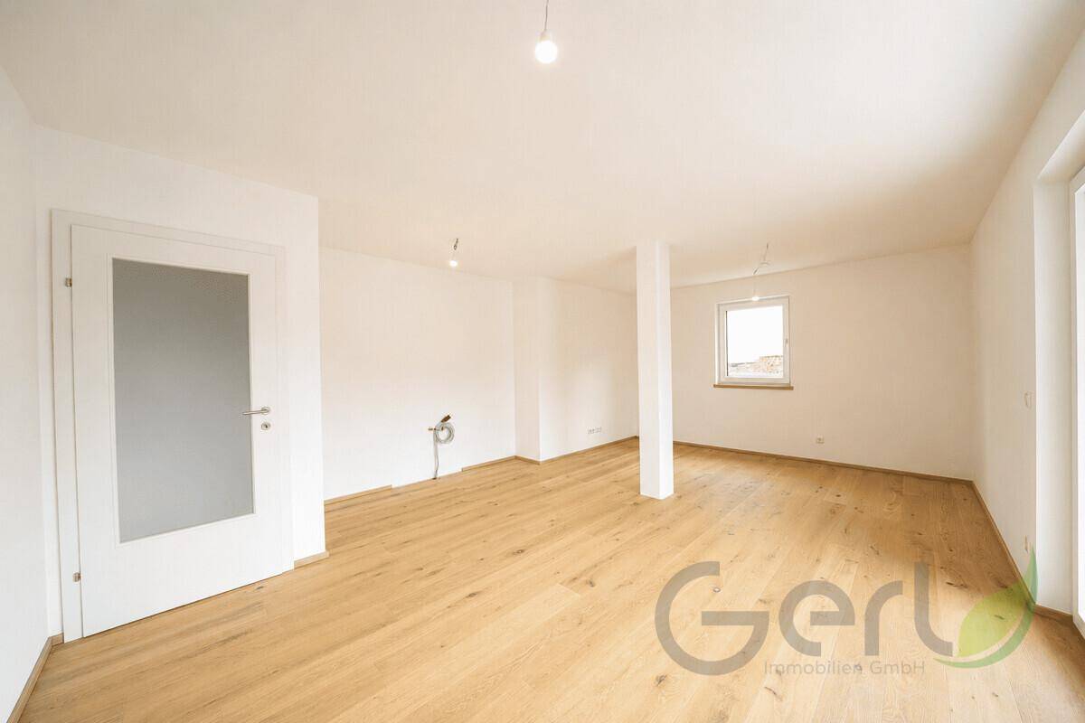 Geräumiges, leeres Wohnzimmer mit hellem Holzboden, großen Fenstern und viel Tageslicht.