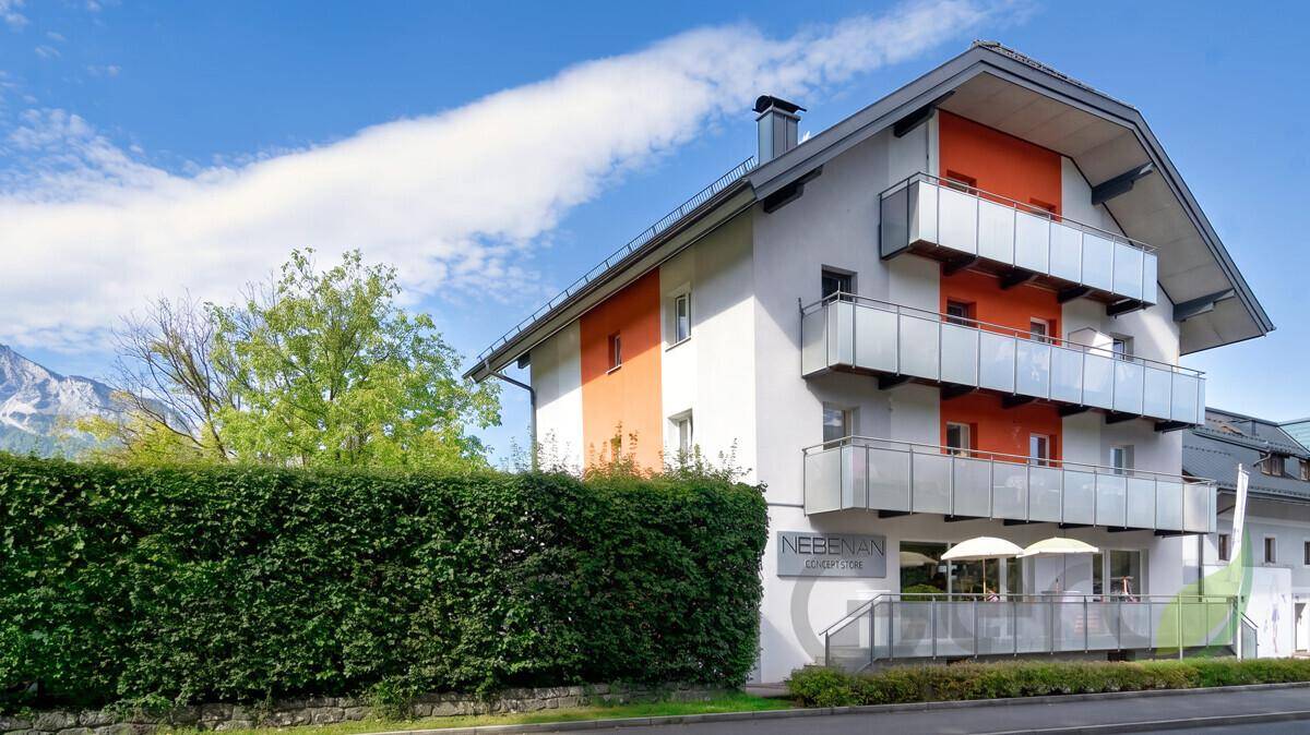 Moderne Gebäudefassade mit mehreren Balkonen, orangefarbenen Akzenten und grüner Hecke.