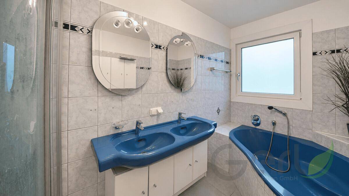 Badezimmer mit blauen Sanitärobjekten, Doppelwaschbecken, Badewanne und hellen Fliesen.