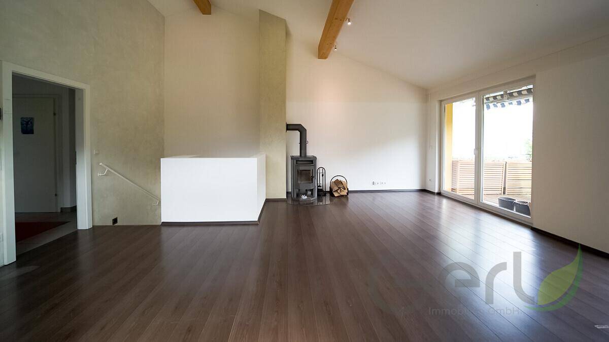Geräumiger, leerer Wohn-Essbereich mit dunklem Holzboden, Kaminofen und Balkonzugang.