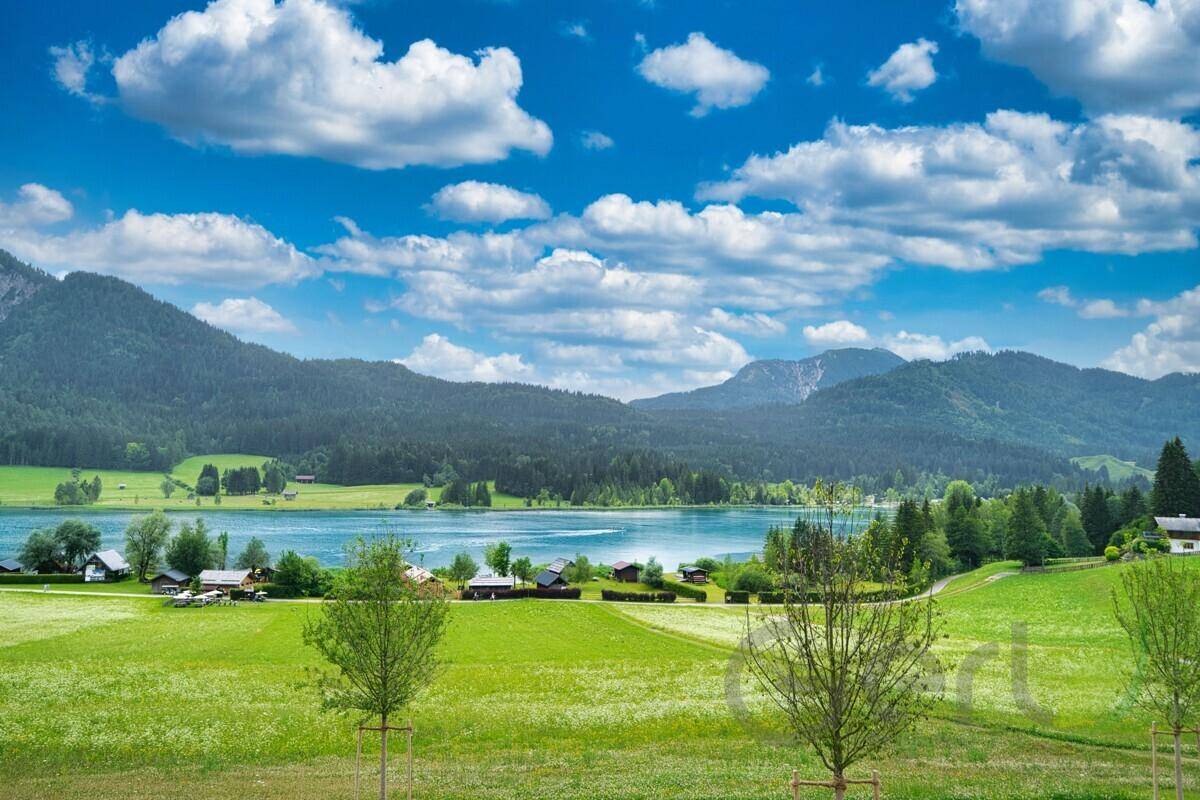Malerischer Blick auf einen klaren Bergsee, umgeben von grünen Hügeln und Wäldern unter blauem Himmel.