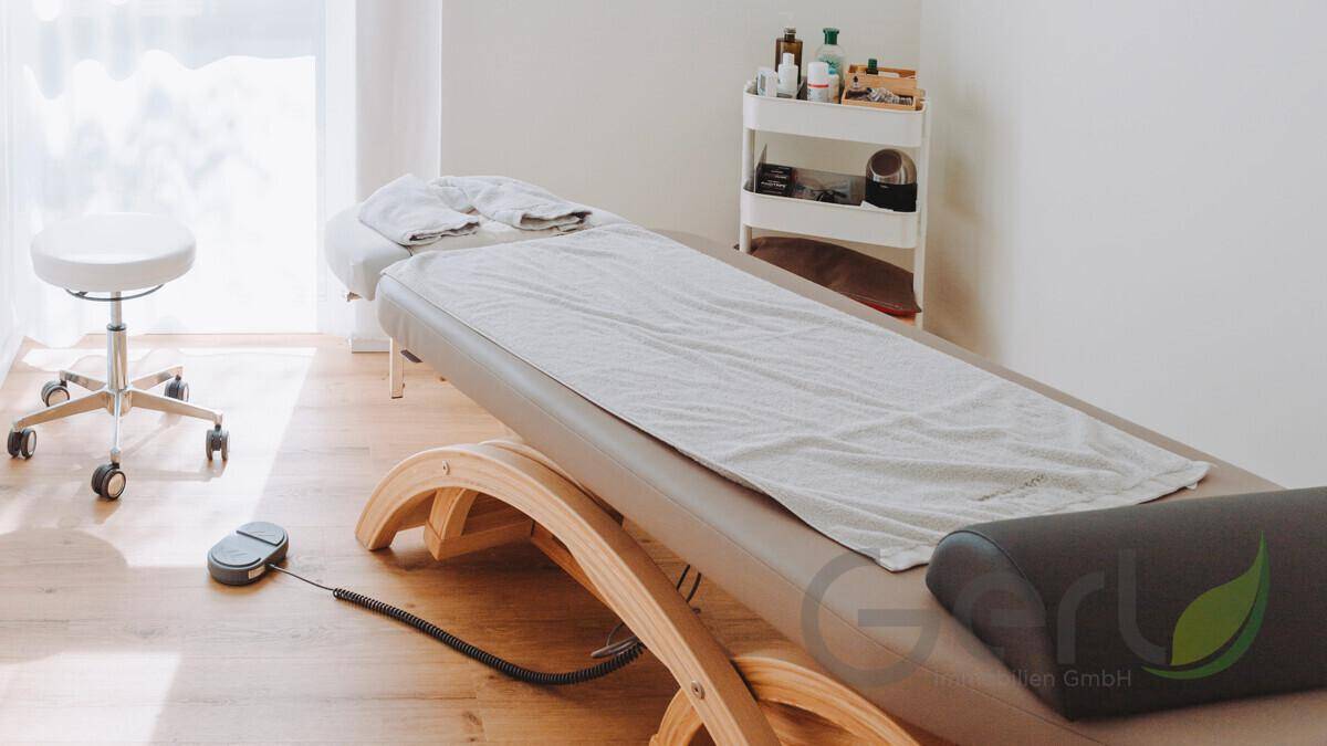 Behandlungsraum mit einer weiteren Massageliege, Rollhocker und einem Servierwagen für Utensilien auf Holzboden.