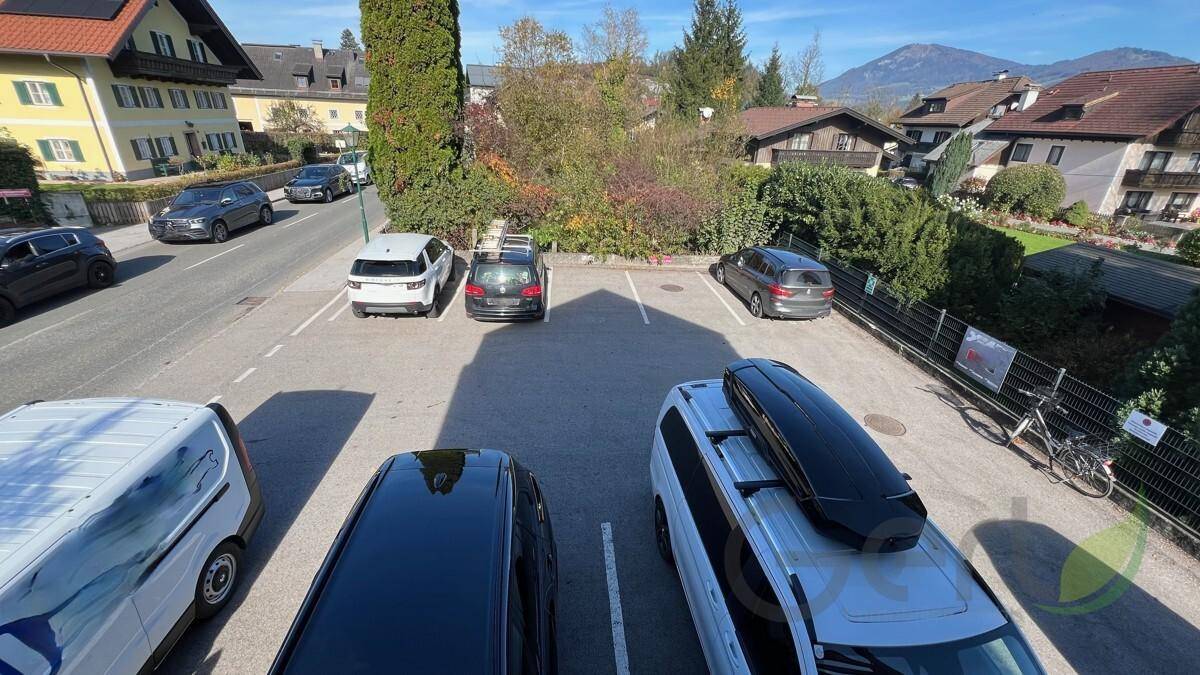 Parkplatz mit mehreren Fahrzeugen, umgeben von Wohnhäusern und Bäumen, mit Bergblick im Hintergrund.