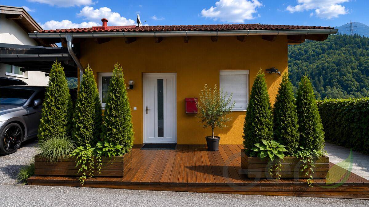 Gelbes Einfamilienhaus mit Holzterrasse, grünen Pflanzen und gepflegtem Vorgarten.