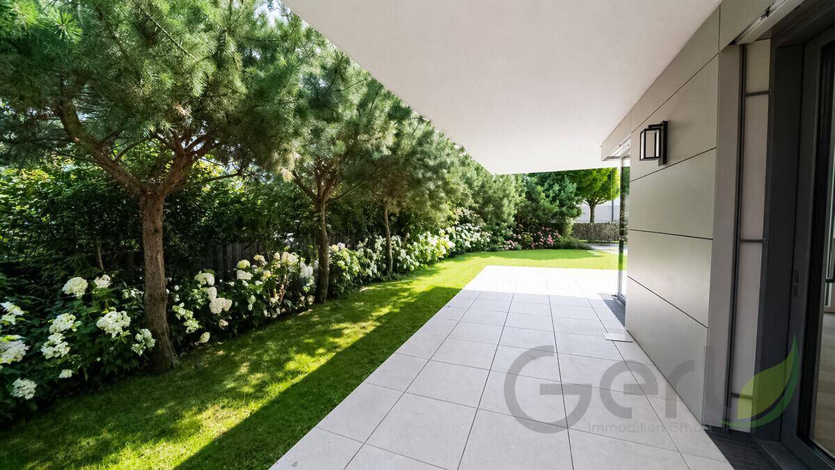 Sonniger Garten mit Rasen, Bäumen und einer gefliesten Terrasse entlang des Gebäudes.