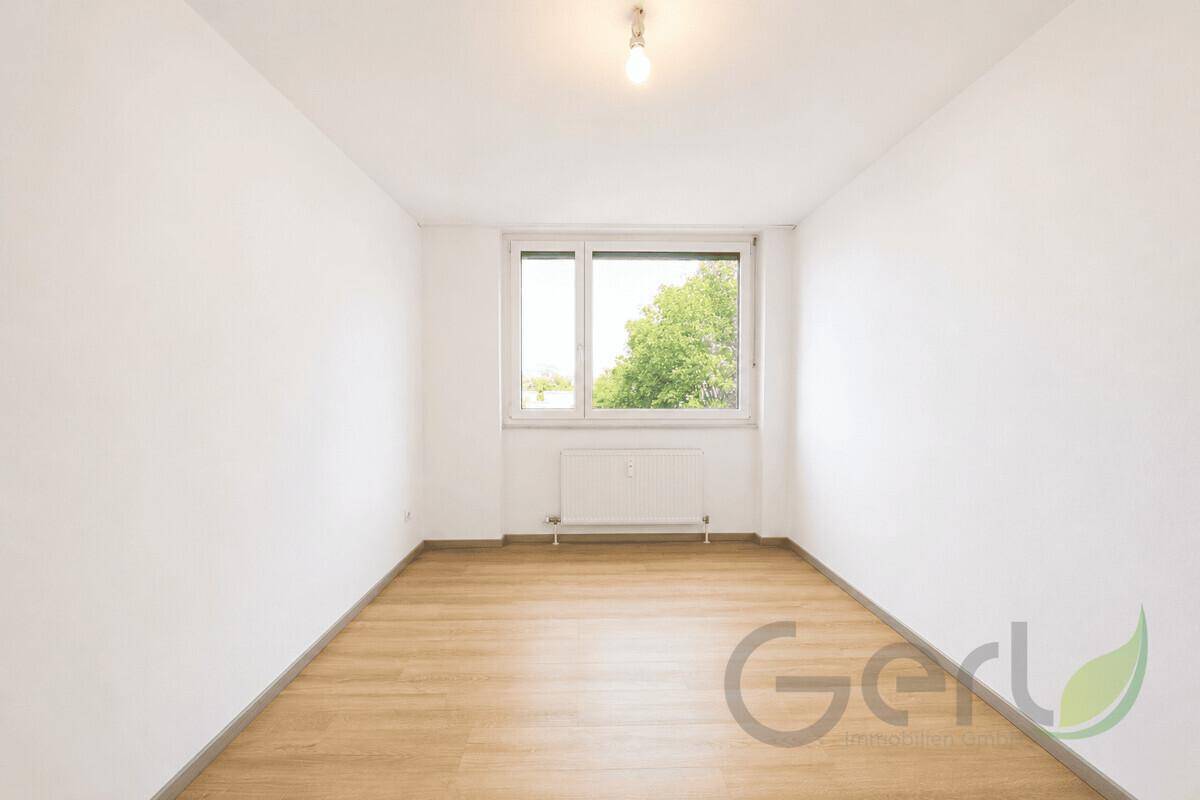 Leeres Schlafzimmer mit Laminatboden, weißen Wänden und Blick ins Grüne durch das Fenster.