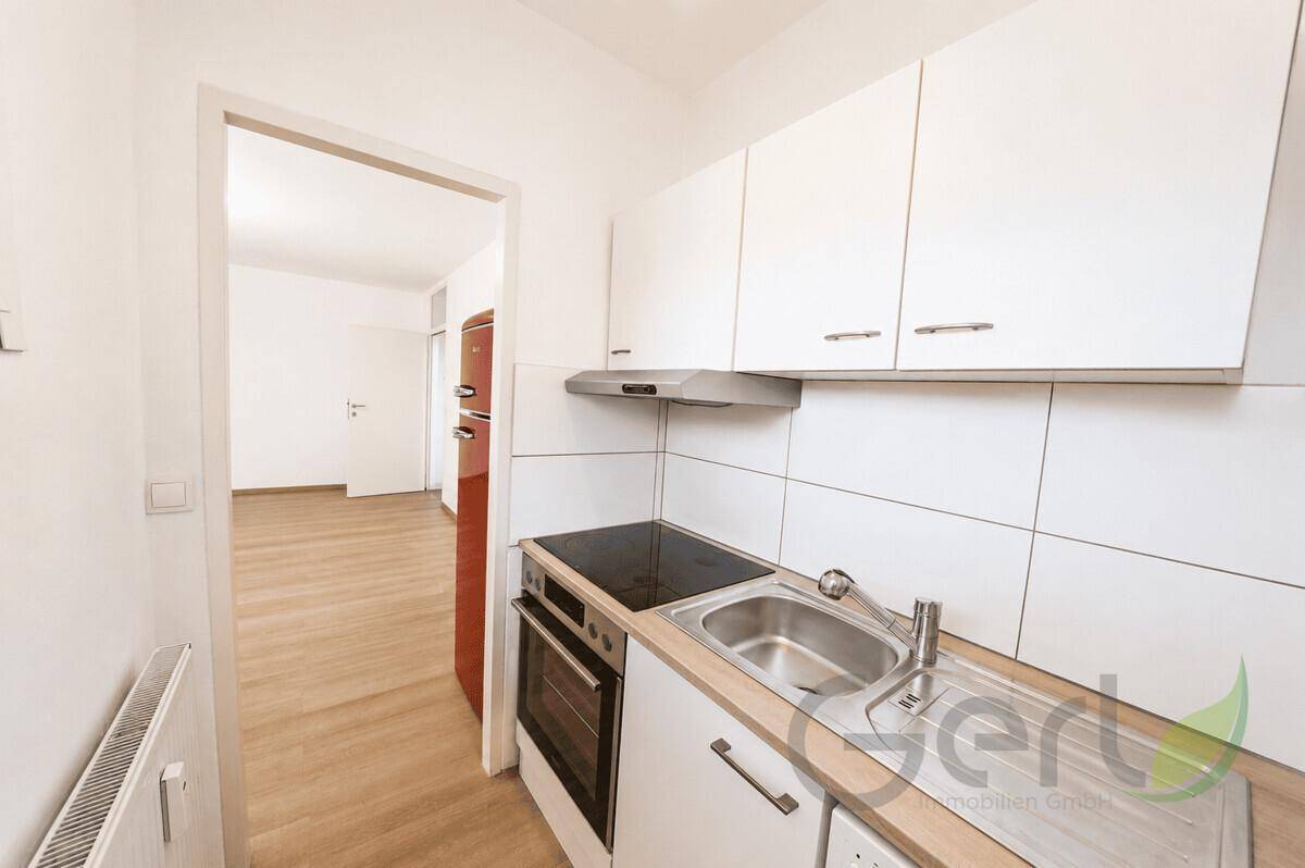 Kleine Einbauküche mit weißen Schränken, Kochfeld und Spüle, offen zum Wohnbereich.