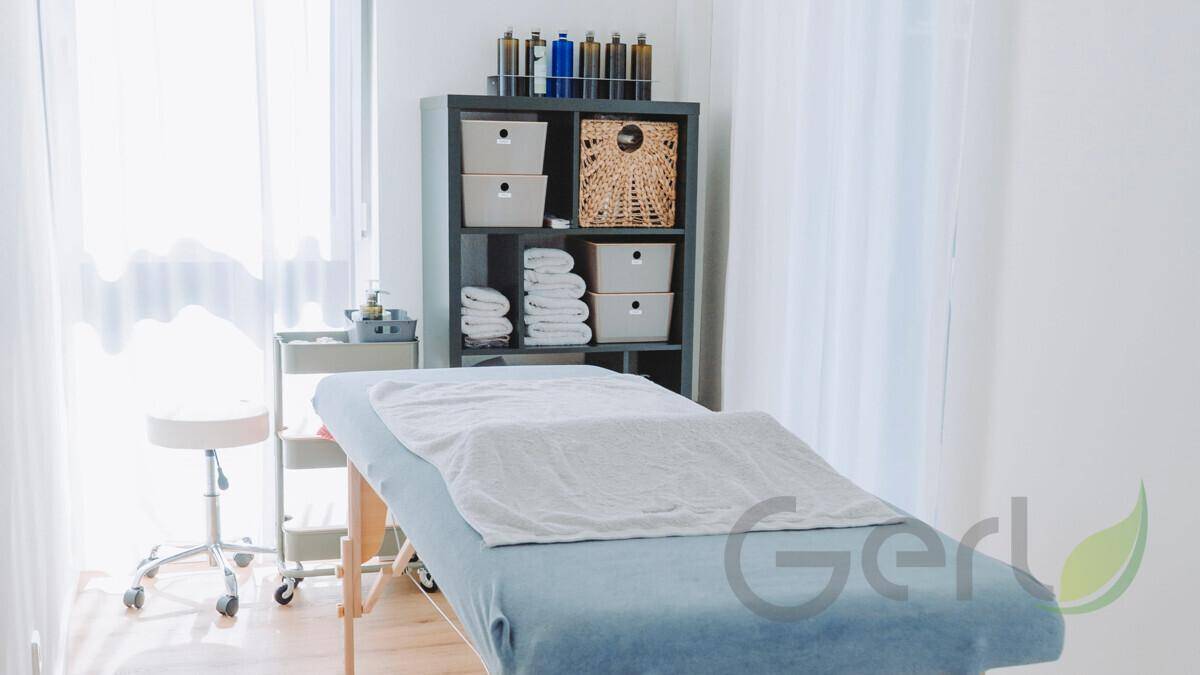 Heller Behandlungsraum mit Massageliege, Regalsystem und viel natürlichem Licht durch die Fenster.