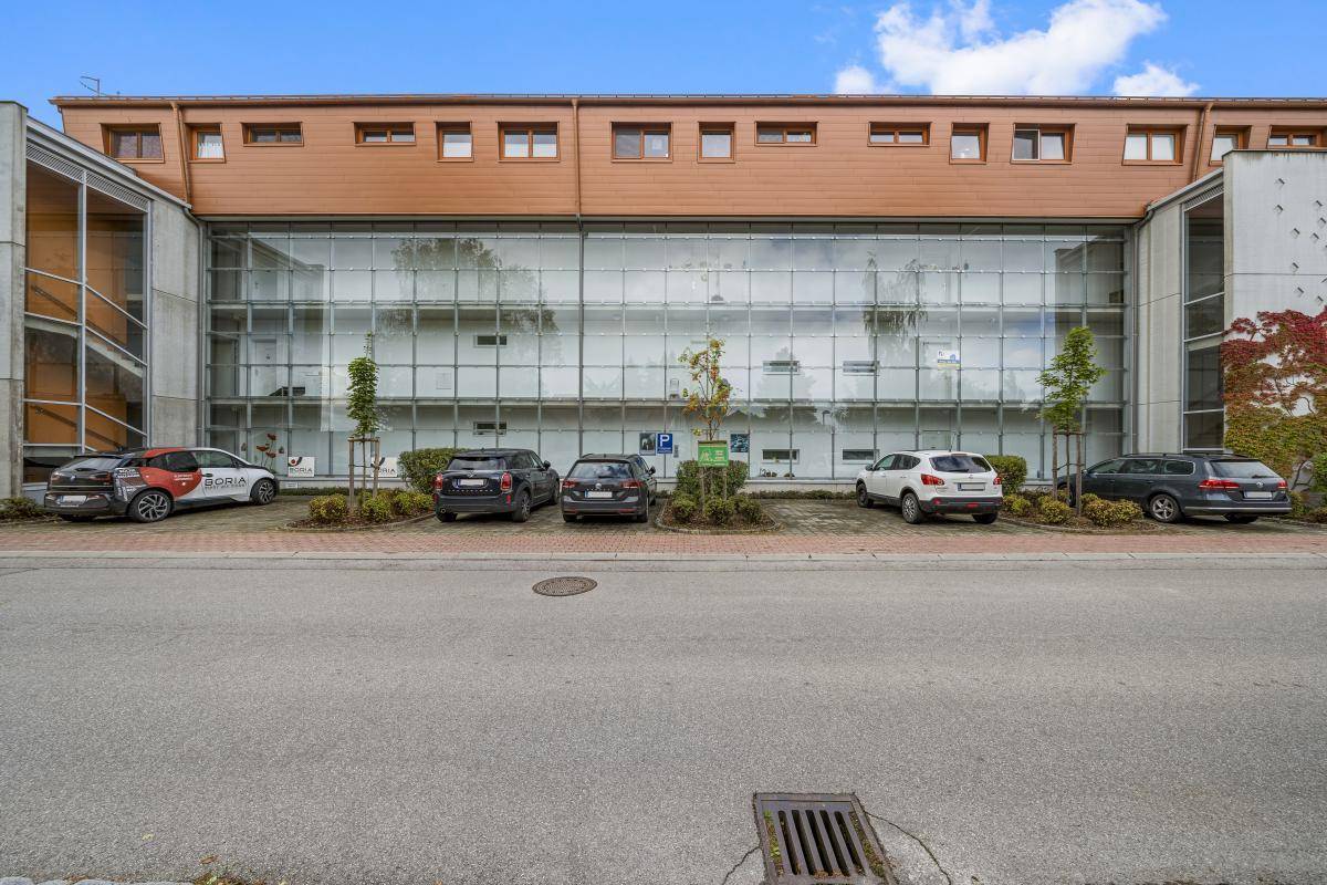Frontansicht des Gebäudes mit großer Glasfassade und davor liegenden Parkplätzen für Fahrzeuge.