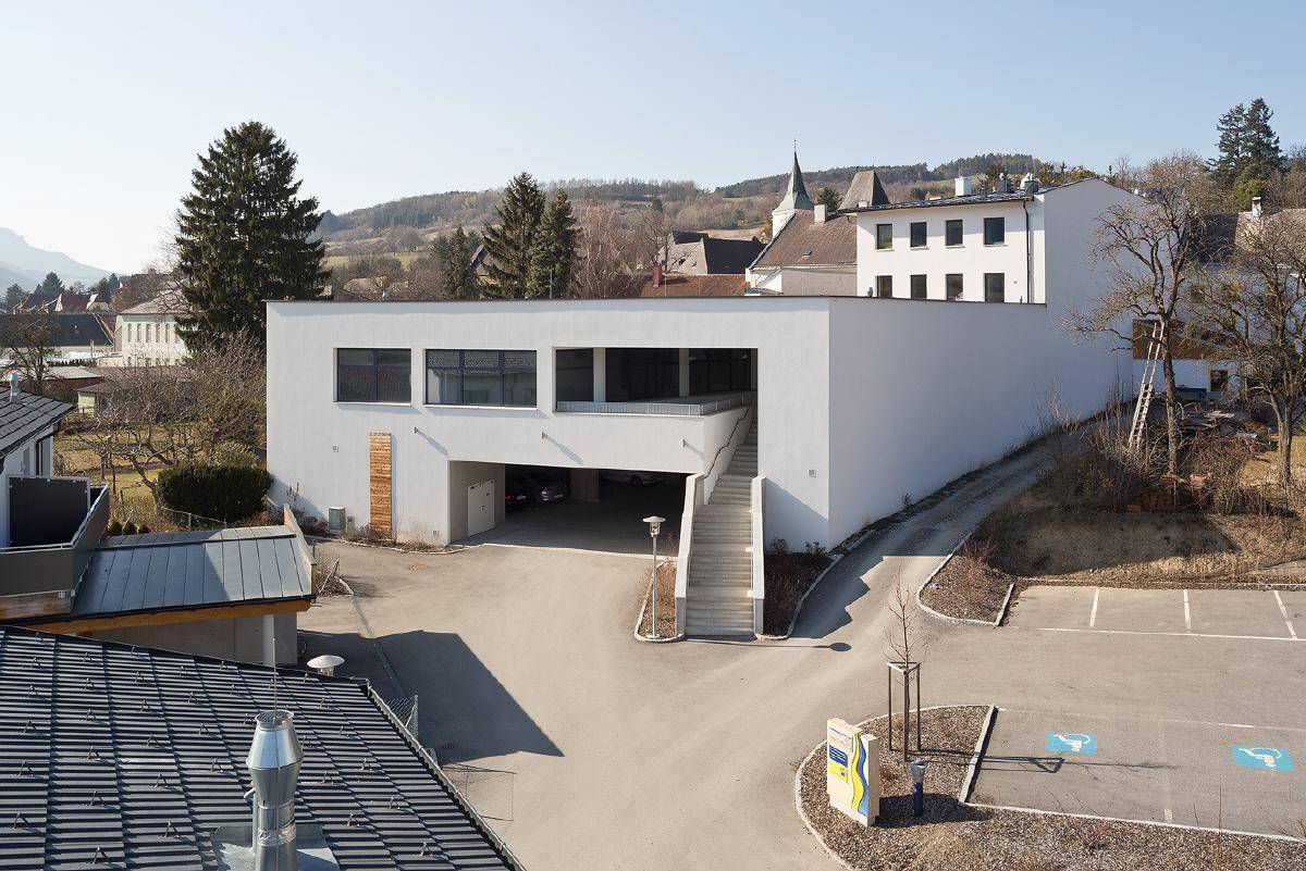 Modernes Gebäude mit Flachdach und Parkplätzen in ländlicher Umgebung mit Hügeln und Kirche.