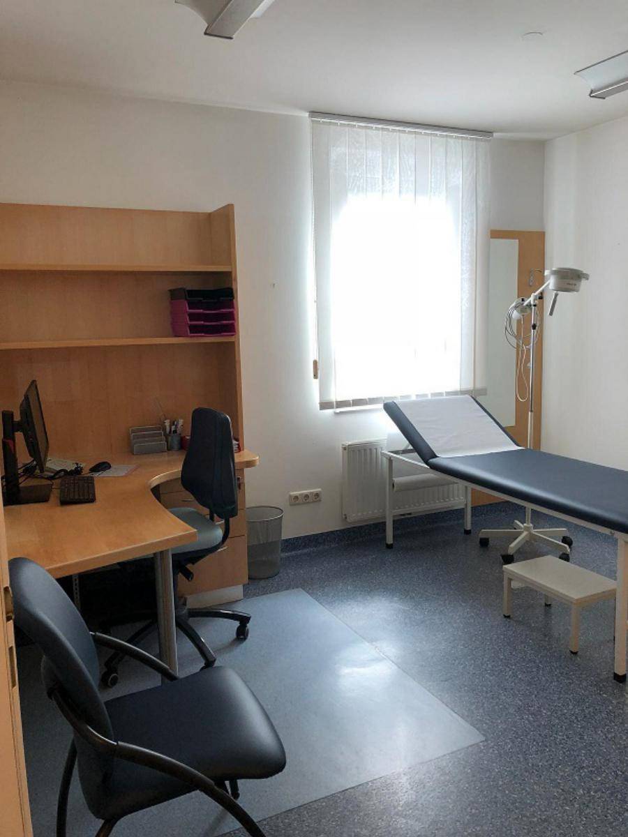 Untersuchungszimmer mit Liege, Schreibtisch und Fenster, ausgestattet für medizinische Zwecke.