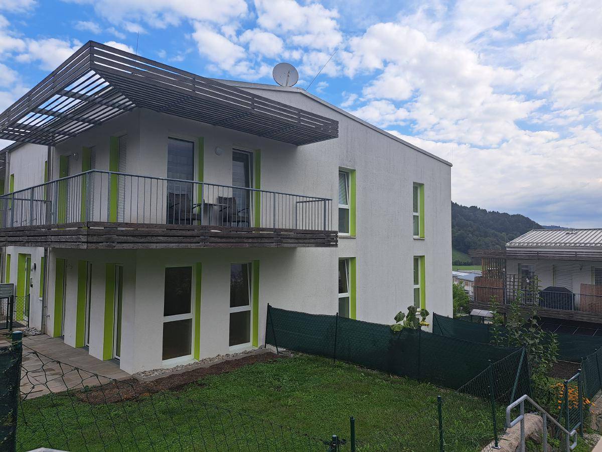 Moderne Wohnanlage mit weißer Fassade, grünen Fensterrahmen und Balkonen mit Holzpergolen vor einer Hügellandschaft.
