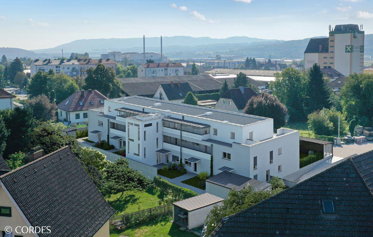 Luftaufnahme eines modernen weißen Gebäudes in einer Wohngegend mit Industrieanlagen und Hügeln im Hintergrund.