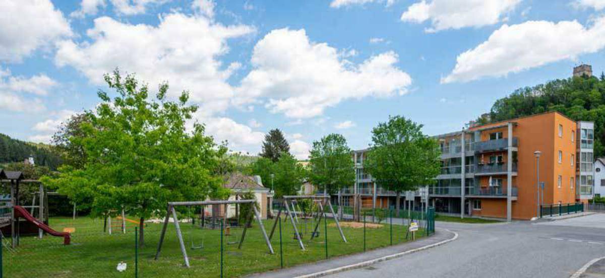Grüner Spielplatz mit Schaukeln und Rutsche vor dem modernen Wohngebäude mit Bergblick.