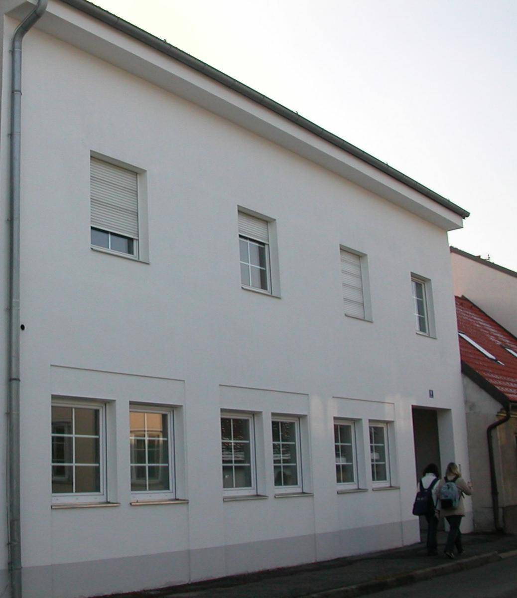Seitenansicht eines gepflegten Gebäudes mit weißer Fassade und mehreren Fenstern, teilweise mit Rollläden.