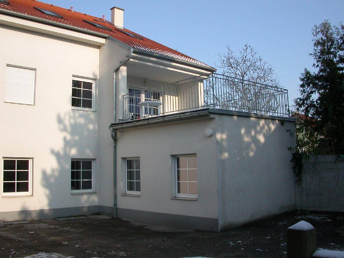 Ansicht der Gebäuderückseite mit einem Balkon und einem roten Ziegeldach unter klarem Himmel.