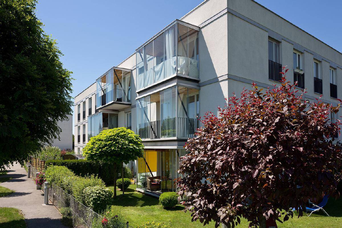 Modernes Wohngebäude mit großen Glasbalkonen und gepflegter Grünanlage unter blauem Himmel.