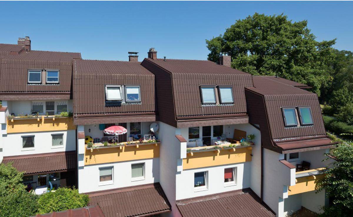 Gebäudekomplex mit braunen Dächern und gelben Balkonen, umgeben von Bäumen an einem sonnigen Tag.