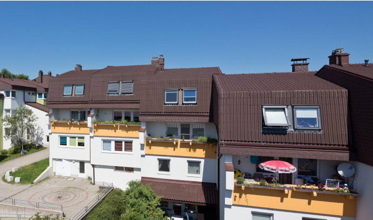 Luftaufnahme eines Mehrfamilienhauses mit braunem Dach und gelben Balkonen unter blauem Himmel.