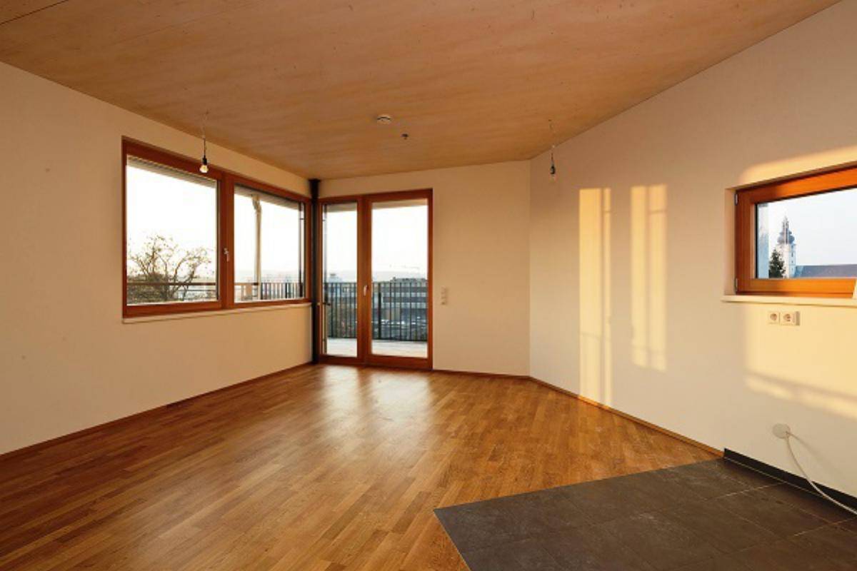 Heller Wohnraum mit Holzboden, großen Fenstern und Zugang zu einem Balkon, durchflutet von Sonnenlicht.