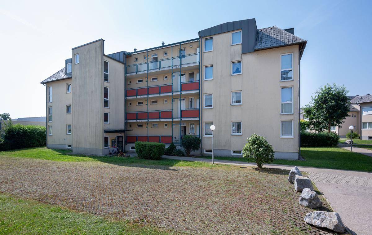 Seitenansicht eines Wohnhauses mit mehreren Balkonen und einer Mischung aus Rasen- und gepflasterten Flächen.