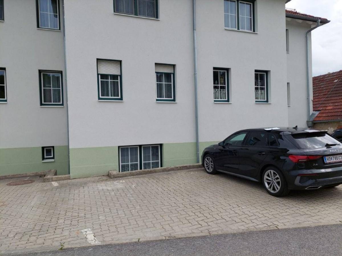 Teilansicht der Gebäudefassade mit einem geparkten Auto und einem gepflasterten Parkplatzbereich.