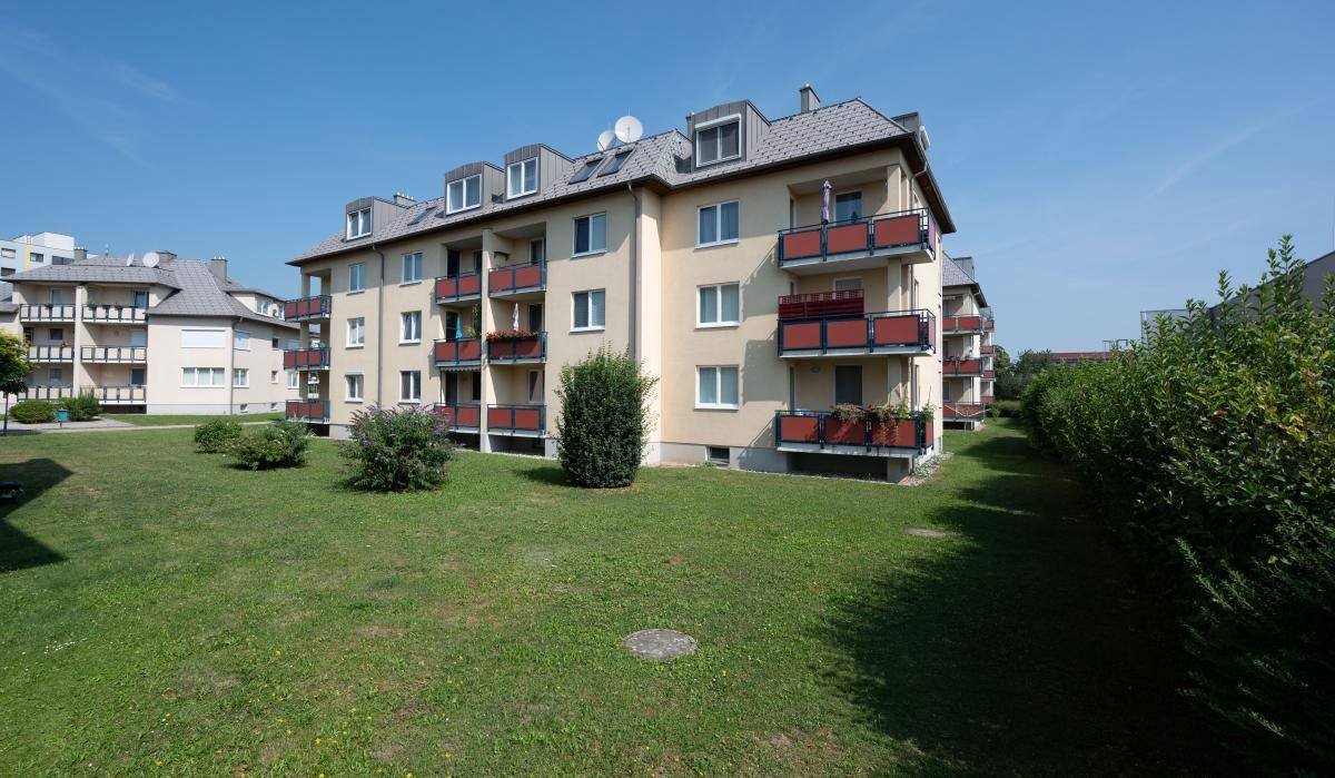 Wohngebäude mit Balkonen und weitläufiger Rasenfläche unter blauem Himmel, umgeben von Hecken.