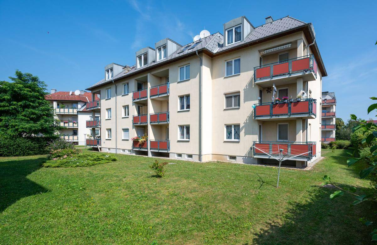 Wohnkomplex mit beiger Fassade, roten Balkonen und Dachgauben, eingebettet in eine sonnige Grünanlage.