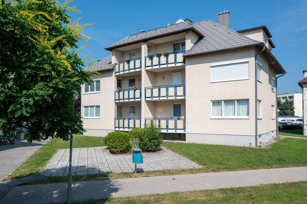Ansicht eines Mehrfamilienhauses mit blauen Balkongeländern und Grünflächen, inklusive Müllbehälter.