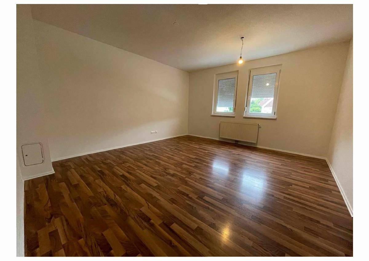 Helles, leeres Zimmer mit warmem Holzboden und zwei Fenstern mit Rollläden.