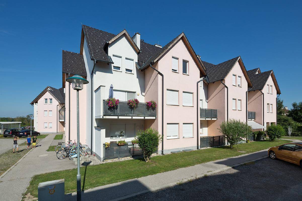 Wohnanlage mit mehreren Reihenhäusern in Pastellfarben, Balkonen und gepflegten Grünflächen an einem sonnigen Tag.