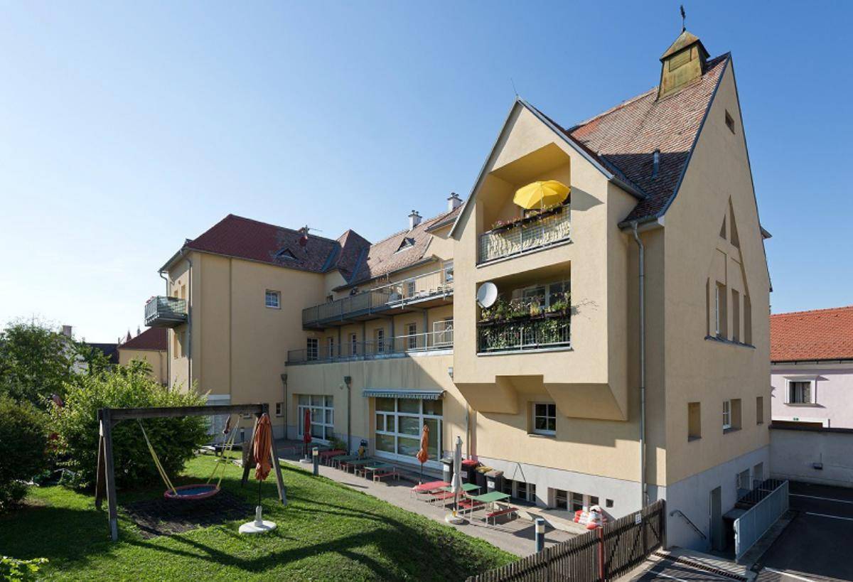 Gesamtansicht des Gebäudes mit Spielplatz, Terrasse und Balkonen an einem sonnigen Tag.