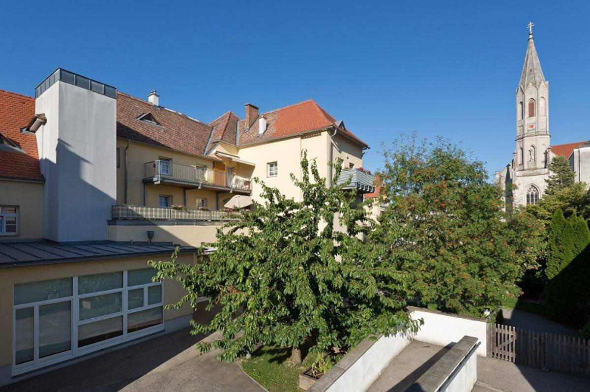 Gebäudeansicht mit Bäumen, Zufahrt und einem Kirchturm im Hintergrund unter klarem Himmel.
