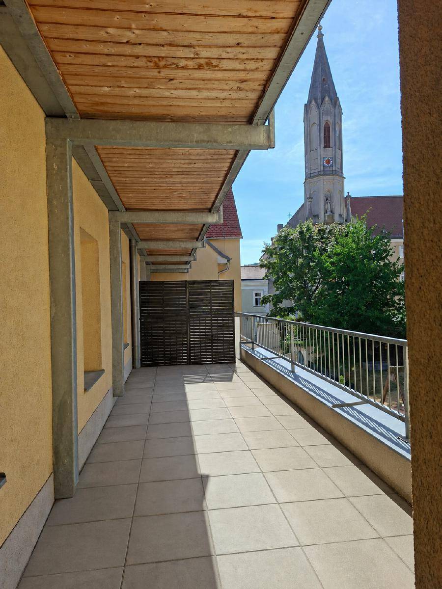 Langer Balkon mit Fliesenboden, Holzdecke und Blick auf einen Kirchturm.