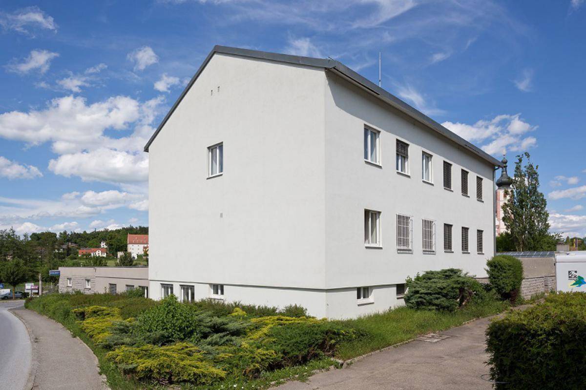 Seitenansicht des hellen Gebäudes mit gepflegter Grünfläche und einer Straße im Vordergrund unter blauem Himmel.
