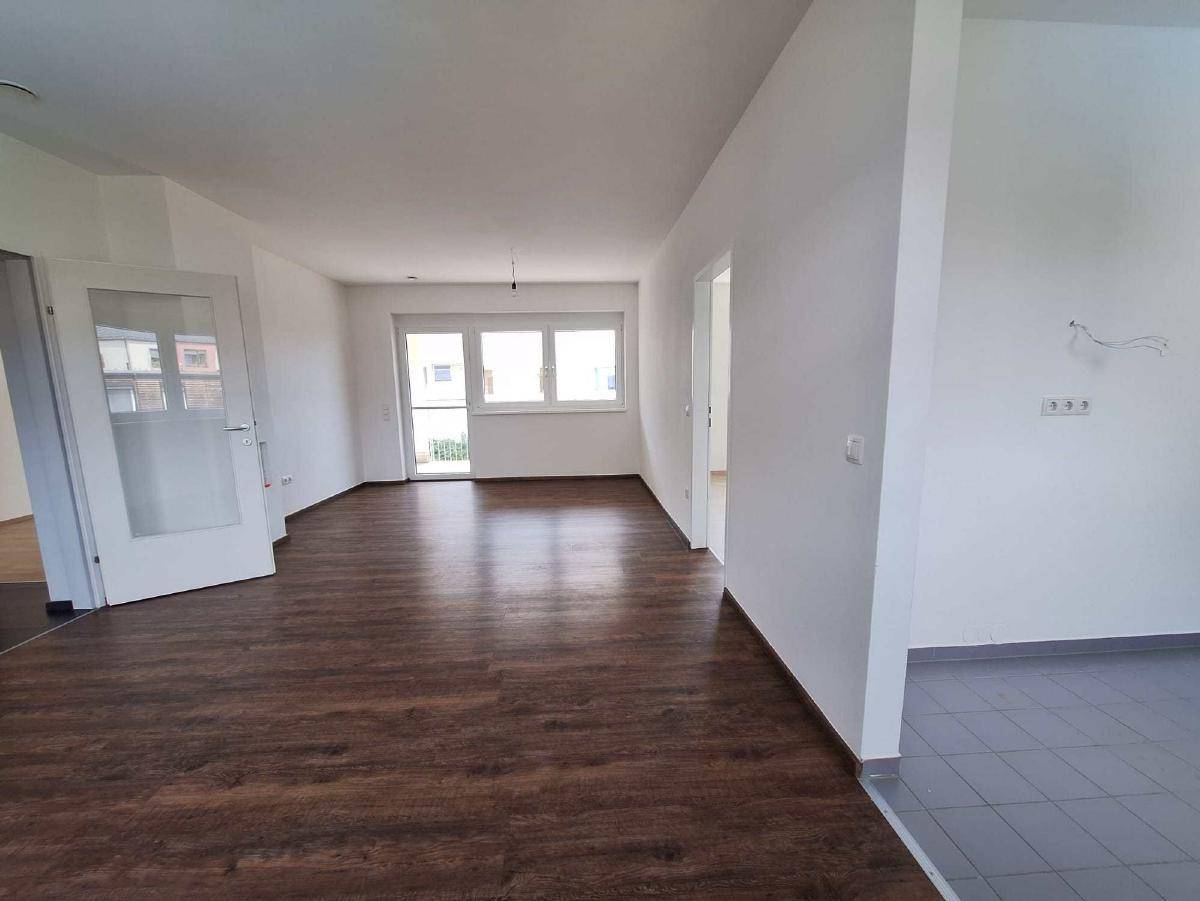 Heller, leerer Wohnbereich mit dunklem Laminatboden und Zugang zu einem Balkon.