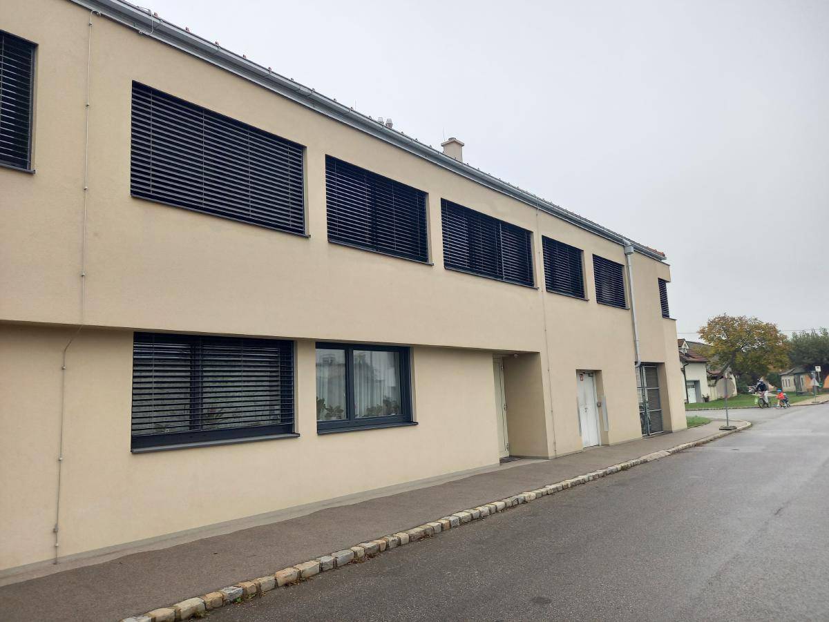 Längsansicht des Gebäudes mit beiger Fassade, vielen Fenstern und geschlossenen Jalousien.