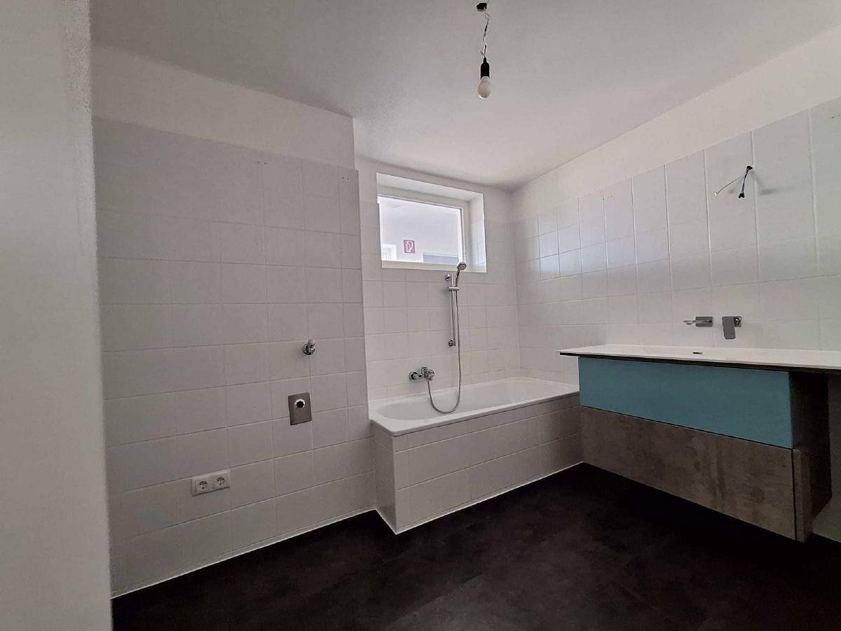 Unmöbliertes Badezimmer mit Badewanne, Fenster und weißen Fliesen an den Wänden.