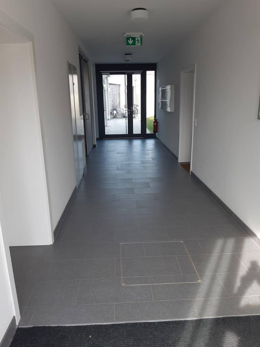 Langer, heller Flur mit grauen Fliesen, weißen Wänden und einer Glastür am Ende.