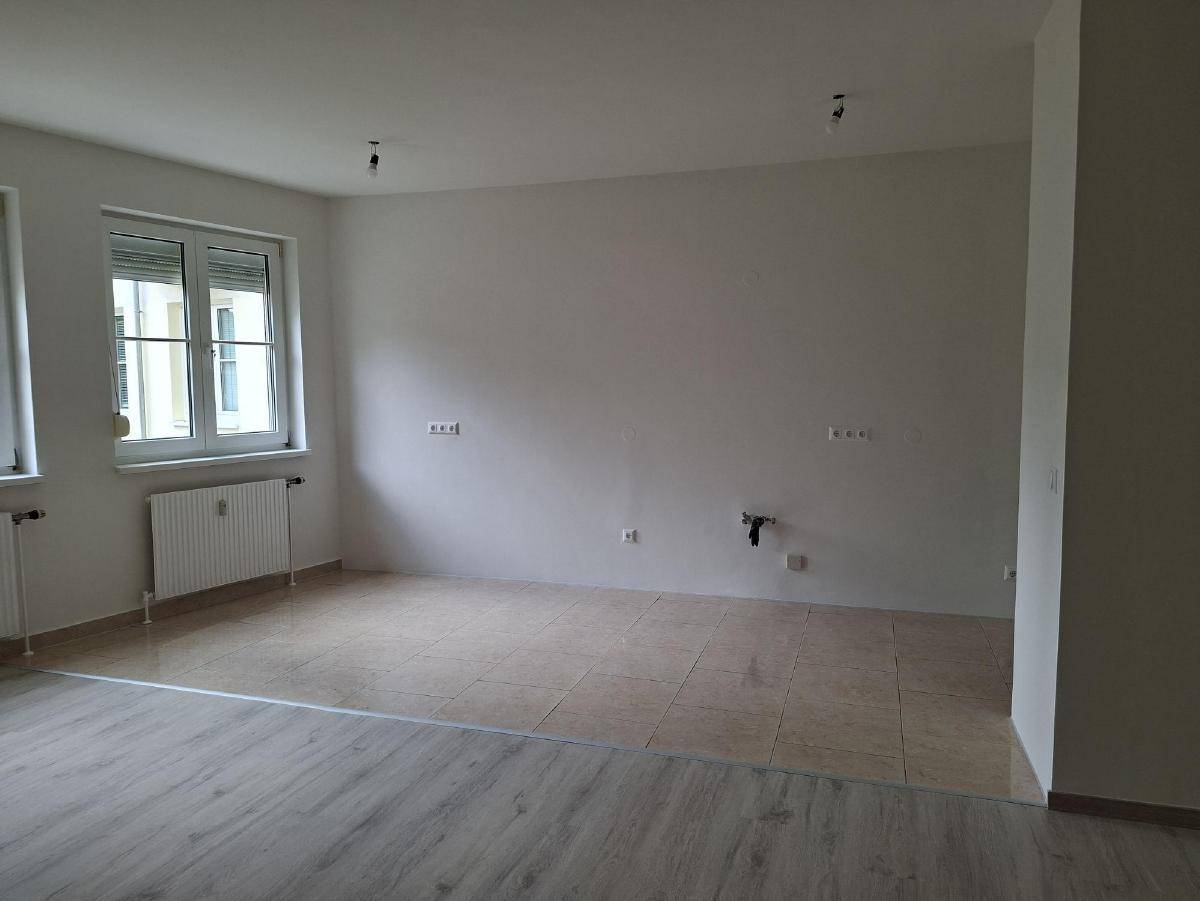 Heller, leerer Raum mit zwei verschiedenen Bodenbelägen und einem Fenster mit Rollladen.
