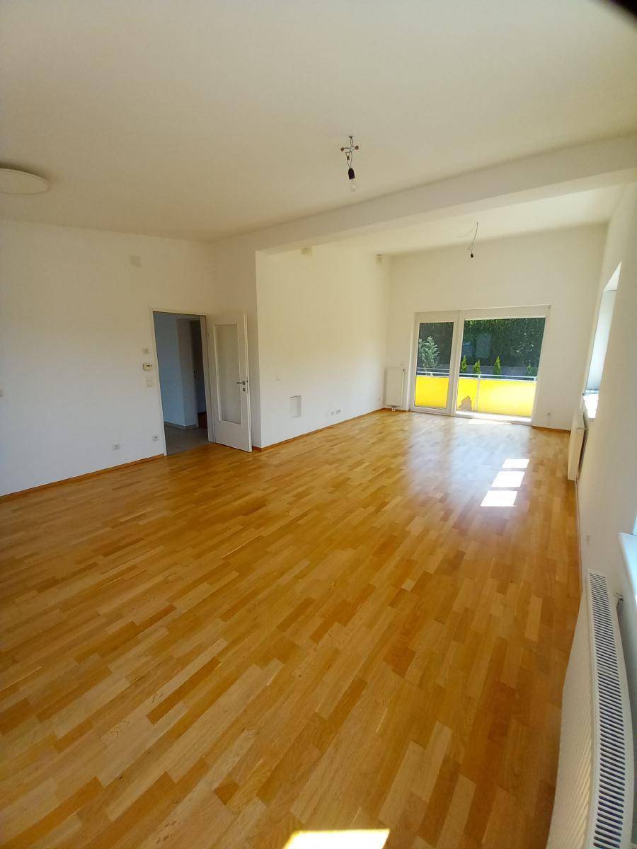 Helles, leeres Wohnzimmer mit Parkettboden und Zugang zu einem Balkon oder einer Terrasse.